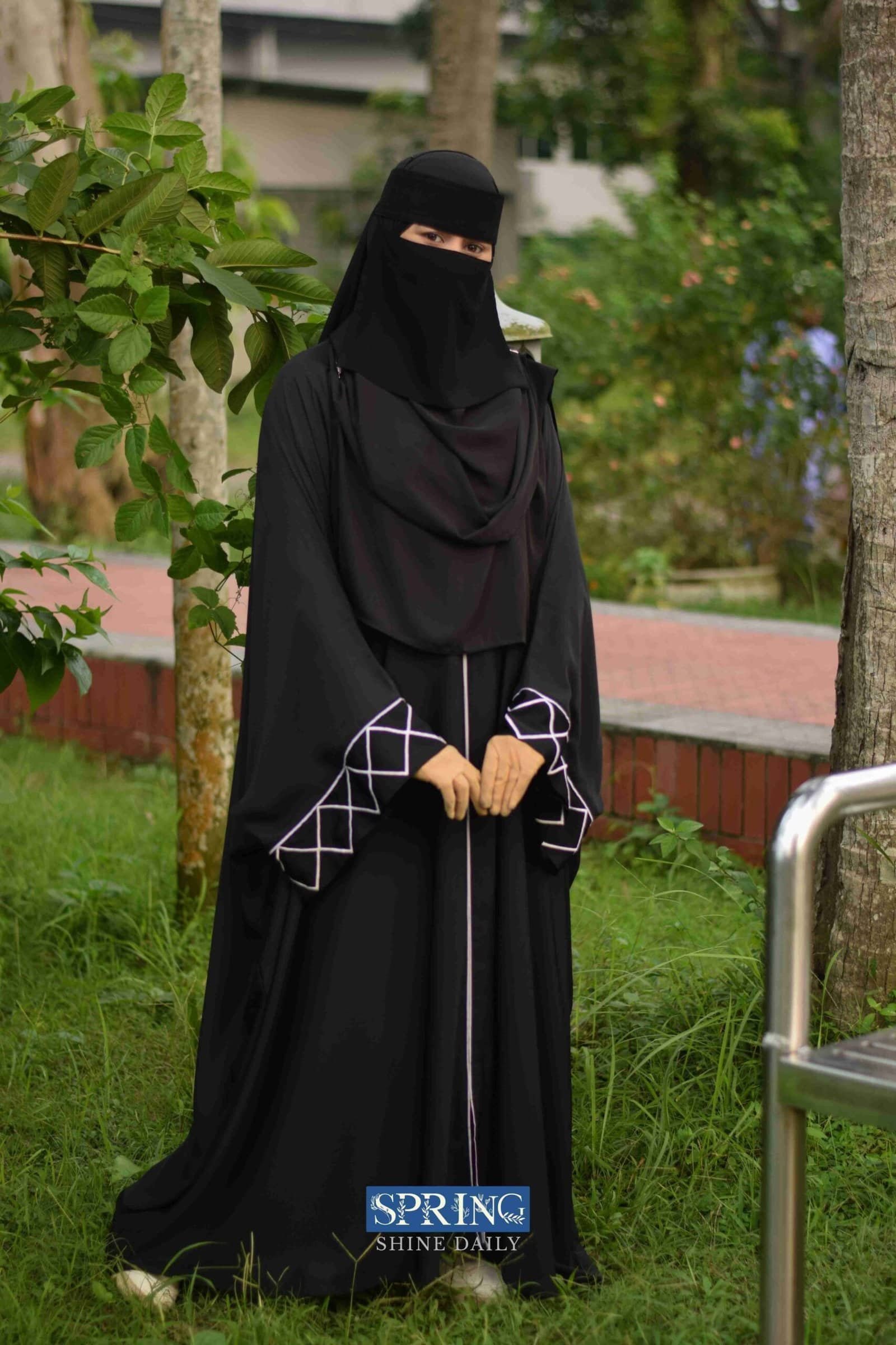 1. Woman in black Islamic abaya and niqab standing outdoors among green trees and grass.