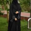Elegant black Islamic abaya with white embroidery details, worn outdoors in a lush green setting.